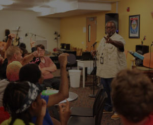 man speaking in front of crowd raising hands in music room
